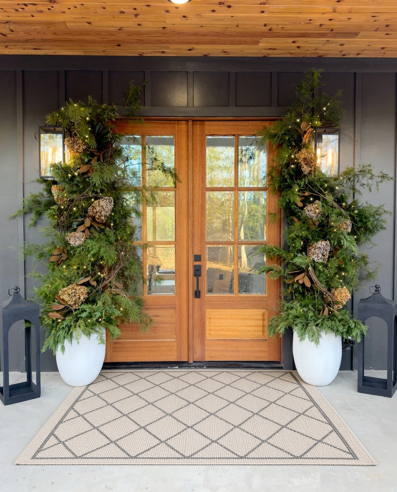 Christmas entrance: evergreen and cedar garlands with dried hydrangea and twinkle lights in white planters at wood double doors