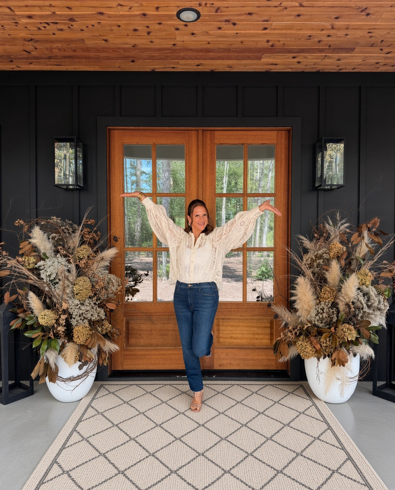 Heidi, arms open, at wood double doors between two tall autumn planters